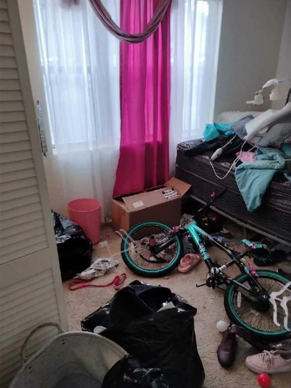 A child's bedroom before cleaning, with a bicycle, bags and clothes covering the floor